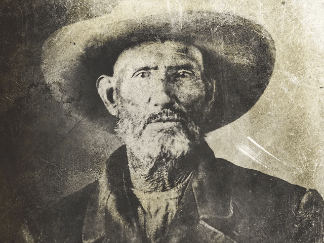 Black and white portrait of an elderly man with a beard and a wide-brimmed hat, wearing a coat and looking directly at the camera.